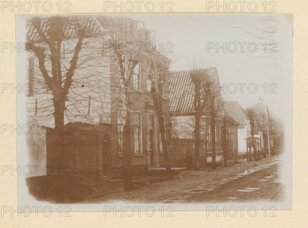 Grootestraat in Batenburg, photograph, page, photographer: Benjamin Charlé (possibly), Batenburg, 1907, paper, pasteboard, baryta paper, daylight collodion silver print, height 150 mm x width 195 mm, street, A sepia-toned street scene showing a row of brick buildings with sloped tiled roofs and tall narrow windows, a line of leafless trees planted along the sidewalk with their bare branches forming a lattice against the sky, a cobblestone road with patches of wetness, an ornate doorway visible on one building, and a small structure or piece of street furniture near the curb further down the street.