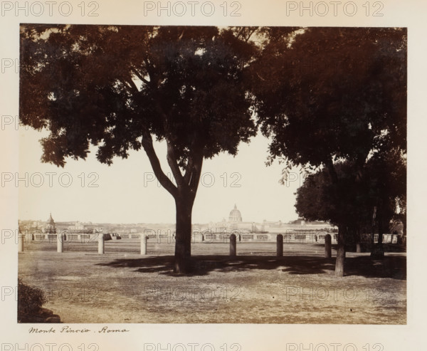 View of Rome from Monte Pincio, Monte Pincio. Rome, album page, photograph, This photo is part of an album., Pincio, 1870 - 1890, photographer, Rome, paper, albumen print, height 202 mm x width 263 mm, garden, city-view in general, 'veduta', trees, public gardens, park, church (exterior), Vatican City, St. Peter's Basilica, A wide, sepia-toned photograph showing a foreground park area with bare, slightly worn ground and shadow patterns, framed by large, leafy trees with dark trunks and dense canopies that create a silhouette against the lighter sky; a row of short stone posts with rounded tops stands between the trees and a balustrade, beyond which an urban riverside panorama is visible with a domed building rising above surrounding structures.