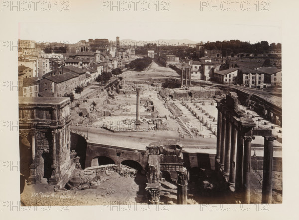 Foro Romano, Forum Romanum in Rome, Italy, album page, photograph, This photo is part of an album., number: '369', Rome, 1870 - 1890, photographer, paper, albumen print, height 188 mm x width 269 mm, market, square, circus, landscape with ruins, Forum Romanum, A wide sepia-toned view of ancient ruins and surrounding buildings showing rows of stone columns and fragmented architectural remains across an excavated area, with several freestanding tall columns visible in the middle ground and a single taller column rising from a rectangular paved area, arched structures and a bridge-like stone construction in the foreground, low multi-story buildings with tiled roofs lining a street on the left, tree-lined paths and sparse vegetation interspersed among the ruins, a range of distant hills along the horizon, and portions of ornate carved stone façades and column capitals visible at the image edges.