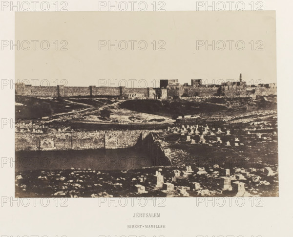 Jerusalem. Birket-Mamillah, Mamilla basin outside the old city wall of Jerusalem, photograph, page, photographer: Auguste Salzmann, Jerusalem, printer (person): Louis-Désiré Blanquart-Evrard, Lille, publisher: Gide et J. Baudry, Paris, 1854 - 1856, printer (person), pasteboard, paper, salted paper print, height 153 mm x width 217 mm, Photo on unnumbered page., city-view in general, 'veduta', pond, pool, Birket-Mamillah, A wide sepia-toned photograph showing a distant stone city wall and clustered buildings along a horizon above a sloping landscape, with a foreground of scattered rectangular stone markers and low ruins, layered terraces and rough ground textures, and a tall narrow tower rising near the right side of the walled structures.