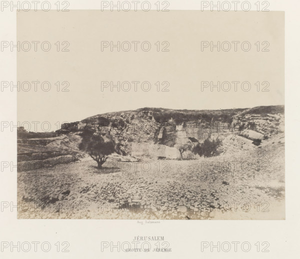 View of the cave of Jeremiah, near Jerusalem, Jérusalem. Grotte de Jérémie, photograph, page, photographer: Auguste Salzmann, Jerusalem, printer (person): Louis-Désiré Blanquart-Evrard, Lille, publisher: Gide et J. Baudry, Paris, 1854 - 1856, printer (person), pasteboard, paper, salted paper print, height 168 mm x width 216 mm, Photo on unnumbered page., cave, grotto, landscape with monument, A sepia-toned photograph showing a rocky landscape with a low cliff or plateau across the midground and a textured foreground of stone and gravel, a few small trees with dark canopies scattered near the base of the cliff, uneven rock formations and ledges along the cliff face, and a large pale sky occupying the upper portion of the image.