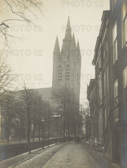 Oude Kerk. (Formerly St. Hypolitus). Tower, View of the Oude Kerk in Delft from the Oude Delft, photograph, inscription: 'Delft. 1st half of the 13th century.', number: '1', inscription: 'The right of reproduction of these photos is reserved by the Publisher.', Delft, c. 1900 - c. 1904, photographer, paper, pasteboard, gelatin silver print, height 155 mm x width 117 mm, parts of church exterior and annexes: tower, Oude Kerk (Delft), A narrow cobblestone street lined with tall buildings on the right and leafless trees on the left leads toward a large church with a central tall spire flanked by four smaller pointed towers and an ornate clock face; a canal with a low railing runs alongside the left side of the street, and a small arched bridge crosses it near the church.