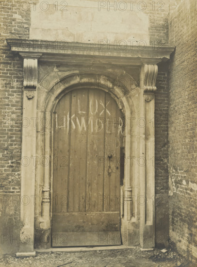 Orphanage. (formerly St. Barbara's Convent.) Oude Vest. Rear exit, Gate of an orphanage on the Westvest in Delft, photograph, number: '10', inscription: 'The right of reproduction of these photos is reserved by the Publisher.', inscription: 'Delft. Early 16th and 17th century.', Delft, c. 1900 - c. 1904, photographer, paper, pasteboard, gelatin silver print, height 156 mm x width 116 mm, gate, entrance, A sepia-toned photograph of a tall arched wooden door set within a stone and brick doorway, with carved stone columns and decorative capitals on either side, a horizontal stone lintel above the arch, weathered vertical wooden planks on the door showing scratches and marks, a round metal knob and a lock plate on the right side of the door, surrounding rough brick walls on the left and right, a stone threshold and worn ground in front of the entrance, and a plain dark border framing the image.