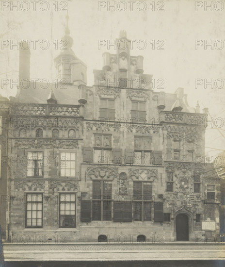Façade of the Gemeenlandshuis on the Oude Delft in Delft, Gemeentelandshuis. Oude Delft WZ No. 167. Front facade, photograph, inscription: 'Delft. Early 16th century.', inscription: 'The right of reproduction of these photos is reserved by the Publisher.', number: '11', Delft, c. 1900 - c. 1904, photographer, paper, pasteboard, gelatin silver print, gelatin silver developing print, height 156 mm x width 116 mm, Gemeenlandshuis, A sepia-toned photograph of an ornate stone building framed by a dark matte border, showing a multi-story façade with decorative carvings, arched windows with shutters and leaded panes, sculpted reliefs above windows and around a central arched doorway, balustraded rooflines, small dormer structures, and a tower with a bulbous finial rising behind the roof; cobbled street and a canal edge appear in front, and bare tree branches are visible at the sides.