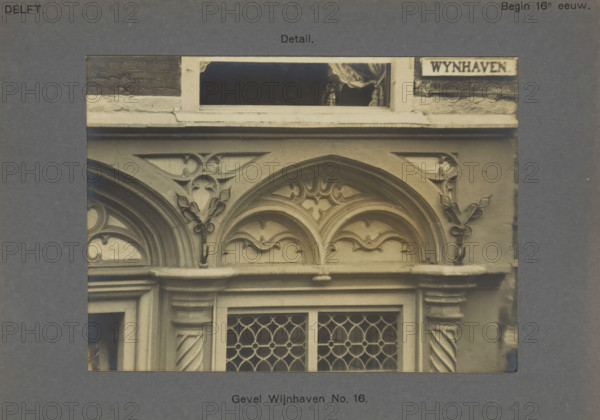 Detail of the decoration of the facade of a building on the Wijnhaven in Delft, Facade Wijnhaven No. 16. Detail, photograph, inscription: 'Delft. Early 16th century.', inscription: 'The right of reproduction of these photos is reserved by the Publisher.', number: '25', Delft, c. 1900 - c. 1904, photographer, paper, pasteboard, gelatin silver print, developed gelatin silver print, height 114 mm x width 156 mm, ornament derived from architectural details, A grayscale photograph showing an ornate building facade with carved stone arches and decorative floral and foliate motifs above a pair of windows with leaded diamond-patterned panes; the upper portion includes a narrow open window with a curtain visible and a rough brick and stone lintel above the decorative frieze.
