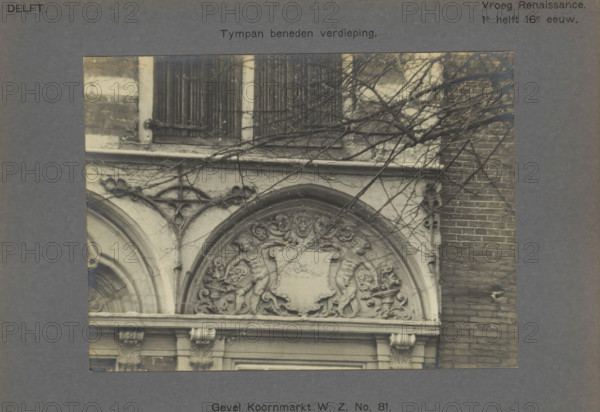 Facade Koornmarkt WZ No. 81. Tympan on the ground floor, Tympanum on the facade of a house on the Koornmarkt in Delft, decorated with two people next to a coat of arms, photograph, inscription: 'The right of reproduction of these photos is reserved by the Publisher.', inscription: 'Delft. Early Renaissance. 1st half of the 16th century.', number: '28', Delft, c. 1900 - c. 1904, photographer, paper, pasteboard, gelatin silver print, developed gelatin silver print, height 115 mm x width 157 mm, urban housing, coat of arms (as symbol of the state, etc.), ornament derived from architectural details, Koornmarkt, A close view of a carved stone facade showing an arched decorative panel with sculpted cherubic figures and ornate floral scrollwork around a central blank shield, framed by curved moldings and supported by carved pilasters with acanthus capitals, with bare tree branches crossing over the stonework and a brick wall visible to the right.