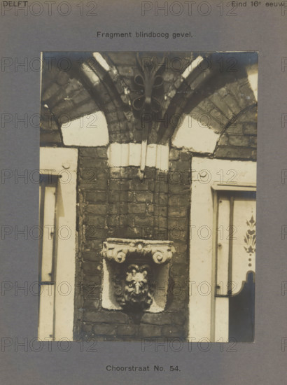 Corb stone decorated with a head in the facade of a building in the Choorstraat in Delft, Choorstraat No. 54. Facade, photograph, inscription: 'The right of reproduction of these photos is reserved by the Publisher.', inscription: 'Delft. Late 16th century.', number: '36', Delft, c. 1900 - c. 1904, photographer, paper, pasteboard, gelatin silver print, developed gelatin silver print, height 157 mm x width 117 mm, urban housing, ornament derived from architectural details, A cropped photograph of a brick wall section showing a decorative stone corbel with a carved face mounted in a recessed square panel, flanked by lighter painted door or window frames on each side; above the corbel the brickwork rises beneath a semi-circular arch with alternating dark and light stone segments and a small carved foliate boss at the arch apex.