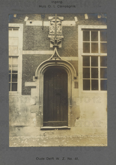 Oude Delft WZ No. 41. Entrance. House of the Dutch East India Company, Entrance to the East India House on Oude Delft in Delft, photograph, inscription: 'The right of reproduction of these photos is reserved by the Publisher.', number: '39', inscription: 'Delft. Early 16th century. Gable stone 1631', Delft, c. 1900 - c. 1904, photographer, paper, pasteboard, gelatin silver print, height 157 mm x width 117 mm, gate, entrance, Oude Delft, A tall arched wooden door set into a stone and brick facade flanked by two tall multi-pane windows, the doorway framed by molded stone with decorative carved elements above including a small ornate plaque and a vertical stone finial, weathered bricks and lighter stone bands surrounding the opening, worn stone steps and a cobbled street in front, and subtle mottling and stains on the lower wall surfaces.