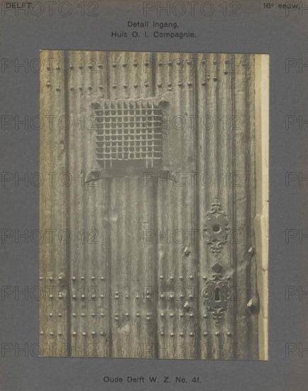 Detail of the entrance to the East India House on Oude Delft in Delft, Oude Delft WZ No. 41. Entrance detail. House OI Company, photograph, inscription: 'The right of reproduction of these photos is reserved by the Publisher.', inscription: 'Delft. 16th century', number: '40', Delft, c. 1900 - c. 1904, photographer, paper, pasteboard, gelatin silver print, height 160 mm x width 120 mm, gate, entrance, lock, door, Oude Delft, A close view of a vertically ribbed wooden door with a square grid-covered opening near the upper center and rows of small rounded studs arranged in horizontal bands, the door surface showing weathered texture and vertical grain. An ornate metal escutcheon plate with a circular hole and decorative flourishes is visible along the right side, and a keyhole plate with an elongated key slot is positioned below it. The door edges show lighter trim on the right and darker worn wood on the left, and the whole image is set against a dark mat background.
