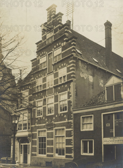 Oude Delft WZ No. 169. Renaissance facade, View of the House of Savoy on the Oude Delft in Delft, photograph, inscription: 'Delft. 17th century. Entrance with bay window of later.', inscription: 'The right of reproduction of these photos is reserved by the Publisher.', number: '43', Delft, c. 1900 - c. 1904, photographer, paper, pasteboard, gelatin silver print, developed gelatin silver print, height 160 mm x width 117 mm, urban housing, decorated gable, Oude Delft, A sepia-toned photograph of a narrow multi-story brick building with a stepped gable and decorative light-colored horizontal stone bands, showing multiple rectangular windows with shutters and panes, a small ornate doorway with a curved pediment, a street lamp in front, an adjacent building with a sloped tiled roof and a tall brick chimney, bare tree branches visible to the left, and a framed museum-style matte surrounding the photograph.