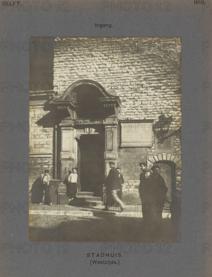 Entrance to the town hall in Delft with boys in front of the gate, Town Hall (West side.) Entrance, photograph, inscription: 'The right of reproduction of these photos is reserved by the Publisher.', number: '46', inscription: 'Delft. 1618.', Delft, c. 1900 - c. 1904, photographer, paper, pasteboard, gelatin silver print, height 155 mm x width 115 mm, town hall, gate, entrance, A sepia photograph shows a stone building facade with a large arched doorway framed by decorative masonry and a smaller arched opening to the side, people standing and walking on the steps and street in front of the entrance, a rectangular sign mounted on the wall beside the doorway, rough textured brickwork covering the wall above, strong sunlight casting distinct shadows, and a dark border surrounding the image.