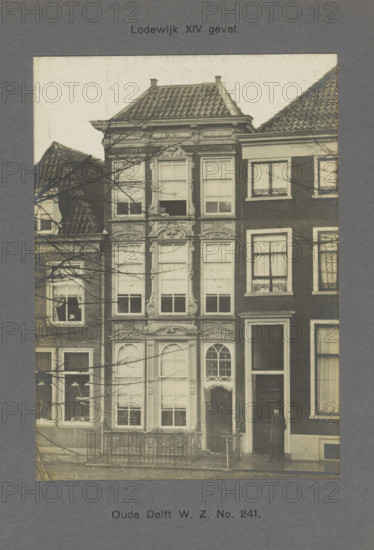View of a building on the Oude Delft in Delft, Oude Delft WZ No. 241. Louis XIV facade, photograph, inscription: 'The right of reproduction of these photos is reserved by the Publisher.', inscription: 'Delft. 1741.', number: '62', Delft, c. 1900 - c. 1904, photographer, paper, pasteboard, gelatin silver print, developed gelatin silver print, height 154 mm x width 110 mm, urban housing, Oude Delft, A sepia photograph of a narrow multi-story building with ornate carved stone window surrounds and decorative panels, flanked by adjacent brick buildings with tiled roofs; the central building has tall sash windows with shutters and a rounded arched doorway with intricate glazing above it, a small fenced area in front, bare tree branches crossing the façade, and a man standing at the entrance of the neighboring building wearing a dark overcoat and hat.