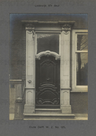 Oude Delft WZ No. 125. Louis XIV door, Door of a building on the Oude Delft in Delft, photograph, inscription: 'Delft. 18th century.', number: '63', inscription: 'The right of reproduction of these photos is reserved by the Publisher.', Delft, c. 1900 - c. 1904, photographer, paper, pasteboard, gelatin silver print, developed gelatin silver print, height 156 mm x width 115 mm, urban housing, door, Oude Delft, A centered ornate wooden door with carved panels and decorative scrollwork set within a pale stone surround featuring fluted pilasters and sculpted capitals, a curved ornamental pediment above a rectangular transom, stone steps leading up to the threshold, a brick wall flanking the doorway on the left, a tall window with a reflection of bare tree branches to the right, and a stone bollard and small vent visible near the base on the left.