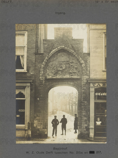Entrance gate to the Bagijnhof in Delft with three people, Bagijnhof. WZ Oude Delft between No. 215a and 217. Entrance, photograph, inscription: 'Delft. 14th to 15th century.', inscription: 'The right of reproduction of these photos is reserved by the Publisher.', number: '8', Delft, c. 1900 - c. 1904, photographer, paper, pasteboard, gelatin silver print, height 155 mm x width 117 mm, alms-house (Dutch: 'hofje'), gate, entrance, Bagijnhof (Delft), A narrow stone archway set into a brick and masonry street facade frames a view down a tree-lined street; the arch has a pointed top with carved relief panels above and textured stonework, flanked by adjacent building fronts with windows and shopfront details, and three men stand silhouetted beneath the arch while a child is visible through a shop window at the right.