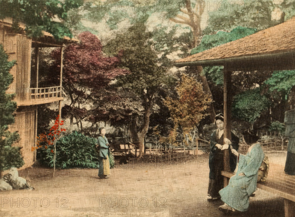 Maple garden at Takinogawa, Three unknown women in a garden, page, photomechanical print, inscription: 'The photograph represents the famous maple garden of Takinogawa, a place of popular resort in the Fall. ...', c. 1891 - in or before 1896, collotype process, height 126 mm x width 173 mm, garden, adult man, Kanto, Tokyo, Kita, Takinogawa, A faded color image shows an outdoor scene with trees and bushes surrounding a wooden structure with a covered porch and a railing, and another roofed area supported by a post. Two women in traditional clothing are near the porch, one seated and one standing, and a person in traditional attire stands farther back near foliage. The ground appears paved and there is a low wooden fence and garden plants in the background.