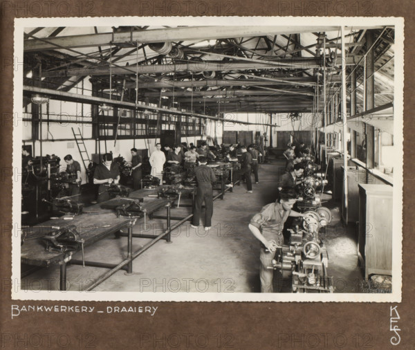 Bench workshop in the Queen Emma School, photograph, View of the bench workshop or turning shop in the Queen Emma School in Surabaya where students are working under the supervision of a teacher. Photo pasted into the photo album presented to JHA van der Hoeven upon the completion of his service in Industrial Education in the Dutch East Indies on July 31, 1933., inscription: 'Bankwerkery of Draaiery, KES', photographer: Fotax (Soerabaja), Surabaya, 1933, Java, East Java, paper, gelatin silver print, height 180 mm x width 246 mm, Queen Emma School, founded in 1912, replacing the Civic Evening School that had existed since 1879., Person, JHA van der Hoeven, vocational education, eg: junior technical school, colonial government, cultural history, education, Dutch East Indies, The, Period, 1911 - 1933, A large workshop interior with long wooden workbenches and rows of metal machines, overhead beams and pulleys spanning the ceiling, and multiple men standing or working at the machines weari...