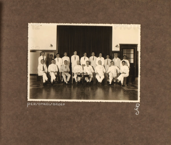 Staff of the Princess Juliana School, photograph, Group photo of the staff of the Princess Juliana School in Yogyakarta. Group of men posing in two rows in the school. Photo pasted into the photo album presented to JHA van der Hoeven upon the completion of his service in Industrial Education in the Dutch East Indies on July 31, 1933., inscription: 'Personnel Group, PJS', Yogyakarta, 1933, photographer, Java, Central Java, paper, gelatin silver print, height 121 mm x width 175 mm, Princess Juliana School in Yogyakarta, founded in 1917, initially intended only as a training program for construction engineers, Person, JHA van der Hoeven, vocational education, eg: junior technical school, anonymous historical persons portrayed in a group, in a group-portrait, colonial government, cultural history, education, Dutch East Indies, The, Period, 1911 - 1933, A sepia photograph shows a group of men posed indoors in front of a dark curtain and light walls with wall-mounted lamps and a doorway with...