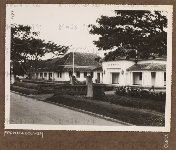 Front buildings of the Queen Emma School, photograph, Front buildings of the Queen Emma School in Surabaya. Photo pasted into the photo album presented to JHA van der Hoeven upon the completion of his service in Industrial Education in the Dutch East Indies on July 31, 1933., inscription: 'Frontgebouwen, KES', photographer: Fotax (Soerabaja), Surabaya, 1933, Java, East Java, paper, gelatin silver print, height 185 mm x width 244 mm, Queen Emma School, founded in 1912, replacing the Civic Evening School that had existed since 1879., Person, JHA van der Hoeven, colonial government, vocational education, eg: junior technical school (building, education, tuition), cultural history, Dutch East Indies, The, Period, 1911 - 1933, A sepia photograph of a low, long building with a tiled roof and a central entrance flanked by columns, set behind a low decorative stone wall and gate posts, with large trees with broad canopies partially obscuring the structure and casting shadows, a paved road in t...