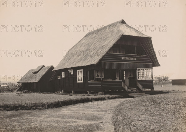 Country house Kaban Djahe, Belawan Ocean Harbor, photograph, View of the country house 'Morgenstond' in Kabandjahe, probably the country house of director Laurens de Groot. Numbered: 230. Part of the group of loose photographs regarding the construction of the port of Belawan on the coast off Medan, Sumatra 1921-1922., date: '3, 10-22', annotation: 'No. 230 Buitenhuis Kaban Djahe', Kabanjahe, 1922-10-03, photographer, Sumatra, paper, gelatin silver print, developing gelatin silver print, height 160 mm x width 220 mm, hut, cabin, lodge, economic history, overseas history, traffic and transport, infrastructure, harbor, dredging, Period, 1921 - 1922, A large wooden house with a steep thatched roof sits on stilts above the ground, featuring a covered porch with steps leading up, shuttered windows with diagonal panels, and a balcony area under the eaves; to the left a smaller thatched structure with a pitched roof is partially visible, and to the right more low buildings can be seen in the ...