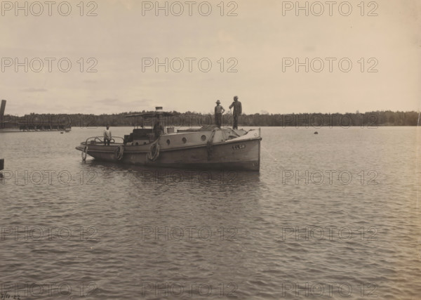 Motor tug 'Elsa', Ocean Harbor Belawan, photograph, The motor tug 'Elsa'. Numbered: 235. Part of the group of individual photographs regarding the construction of the harbor of Belawan on the coast off Medan, Sumatra 1921-1922., annotation: 'No. 235 Motor tug 'Elsa'', date: '7, 10-22', Belawan, 1922-10-07, photographer, Sumatra, paper, gelatin silver print, developing gelatin silver print, height 160 mm x width 220 mm, tug, pusher tug, economic history, overseas history, traffic and transport, infrastructure, harbor, dredging, Period, 1921 - 1922, A small boat floats on calm water with three people standing on its deck; the vessel has a covered central area with a small funnel and round portholes along the hull, coiled ropes draped over the sides, and a lifebuoy hanging at the stern. The boat is positioned near a low treeline that stretches across the background and a wooden pier is visible to one side. The sky is light and mostly clear with a few thin clouds, and the water surface sho...