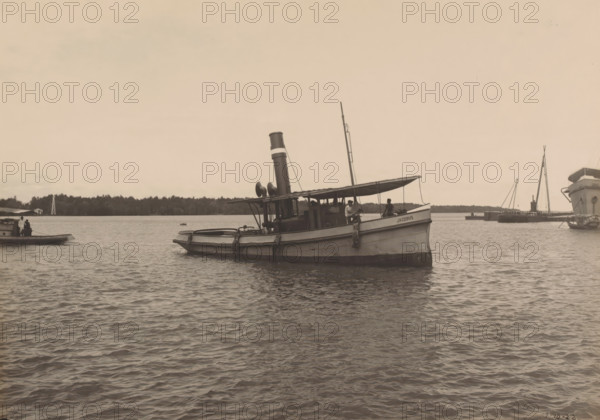Tugboat 'Jacobus', Ocean Harbor Belawan, photograph, The motor tugboat 'Jacobus'. Numbered: 236. Part of the group of individual photographs regarding the construction of the harbor of Belawan on the coast off Medan, Sumatra 1921-1922., annotation: 'No. 236 Tugboat 'Jacobus'', date: '7, 10-22', Belawan, 1922-10-07, photographer, Sumatra, paper, gelatin silver print, developing gelatin silver print, height 160 mm x width 220 mm, tug, pusher tug, economic history, overseas history, traffic and transport, infrastructure, harbor, dredging, Period, 1921 - 1922, A medium-sized steam launch floats on calm water with a canopy over its midsection, a tall funnel rising behind the canopy, and a mast extending upward toward the sky; two people are visible on the deck, one seated near the bow and another near the cabin, and coiled ropes hang along the hull. In the background a tree-lined shore runs across the horizon with several other boats and masts visible near the right edge of the scene, and a...