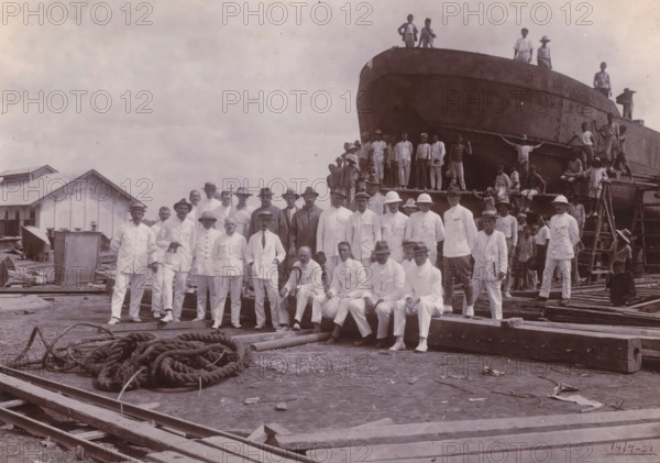 Farewell visit Chief Engineer Fritzlin, Belawan Ocean Port, photograph, Group photo at the construction site in front of the bow of a ship, on the occasion of the farewell visit of Chief Engineer Fritzlin, second director of the port of Belawan, July 14, 1921. In the foreground the Dutch in white uniforms, behind them Indonesian boys on and in front of the ship. Numbered: 47. Part of the group of individual photos regarding the construction of the port of Belawan on the coast off Medan, Sumatra 1921-1922., date: '17, 7-21', inscription: 'No. 47. Ocean Port Belawan, Farewell Visit Chief Engineer Fritzlin', Belawan, 1921-07-14, photographer, Sumatra, paper, daylight gelatin silver print, height 160 mm x width 220 mm, harbor, building-site, economic history, overseas history, traffic and transport, infrastructure, Period, 1921 - 1922, A large group of people pose in front of a partially constructed or repaired large vessel on blocks, with many standing on the ground wearing light-colored ...