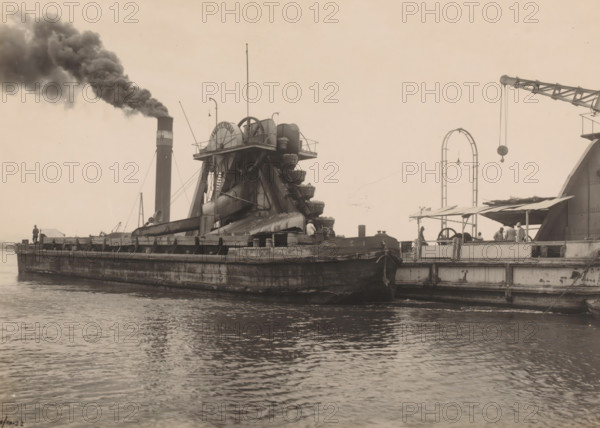 Dredger Amsterdam, Ocean Harbor Belawan, photograph, The dredger 'Amsterdam' on the water next to another ship. Numbered: 240. Part of the group of individual photographs regarding the construction of the harbor of Belawan on the coast off Medan, Sumatra 1921-1922., date: '9, 10-22', inscription: 'No. 240 Baggermolen Amsterdam', Belawan, 1922-10-09, photographer, Sumatra, paper, gelatin silver print, height 160 mm x width 220 mm, building-site, dredge, dredging machine, economic history, overseas history, traffic and transport, infrastructure, harbor, dredging work, Period, 1921 - 1922, A sepia-toned waterfront scene showing a large, flat-topped barge with heavy industrial machinery mounted on it, including a multi-bucket wheel structure with curved chutes and supporting beams, a tall smokestack emitting a dense plume of dark smoke, and various cables and poles rising from the deck. The barge's weathered hull sits low in the water, with visible rails and ladders along its side and a sm...