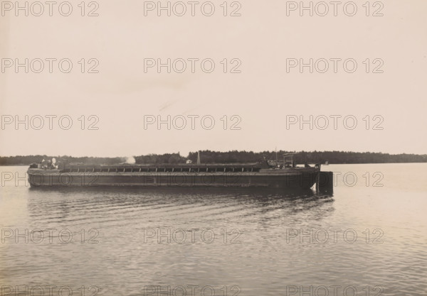 Bottom-dump barge 16, Ocean Harbor Belawan, photograph, Transport ship, bottom-dump barge. Numbered: 255. Part of the group of individual photographs regarding the construction of the port of Belawan on the coast off Medan, Sumatra 1921-1922., date: '12, 10-22', annotation: 'No. 255 Bottom-dump barge 16', Belawan, 1922-10-12, photographer, Sumatra, paper, gelatin silver print, height 160 mm x width 220 mm, building-site, motorized barge, economic history, overseas history, traffic and transport, infrastructure, harbor, dredging, Period, 1921 - 1922, A long, dark-hulled barge sits on calm water with slight ripples, stretching across the middle of the image with a low profile and an open cargo area; a few figures stand near the stern, and small structures and equipment are visible on the deck toward the rear. A tree-lined shoreline runs behind the barge, with scattered taller elements rising above the treeline. The sky is light and mostly clear with faint, diffuse blemishes, and the wate...