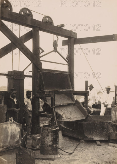 On top of the pouring jetty, Ocean Harbor Belawan, photograph, Scaffolding used to pour the concrete, with workers and supervisors. Numbered: 269. Part of the group of individual photographs regarding the construction of the port of Belawan on the coast off Medan, Sumatra 1921-1922., annotation: 'No. 269 On top of the dumping pier', date: '25, 10-22', Belawan, 1922-10-25, photographer, Sumatra, paper, gelatin silver print, gelatin silver developing print, height 220 mm x width 160 mm, building-site, making the overground structures (, building activities), economic history, overseas history, traffic and transport, infrastructure, harbor, dredging, Period, 1921 - 1922, A large wooden industrial structure with pulleys and ropes dominates the scene, showing a chute or hopper with layers of rough material cascading into a basin below, with wooden beams and diagonal braces forming a frame around it; several metal buckets and cylindrical containers sit on the ground in front of the machinery...