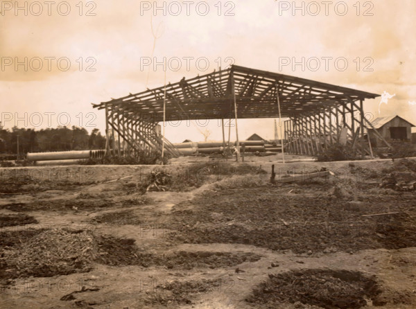 Construction of a shed, photograph, Framework of a shed under construction. Part of the group of individual photographs regarding the construction of the port of Belawan on the coast off Medan, Sumatra 1921-1922., Belawan, 1921 - 1922, photographer, Sumatra, paper, gelatin silver print, height 125 mm x width 170 mm, building-site, making the overground structures (, building activities), economic history, overseas history, traffic and transport, infrastructure, harbor, dredging, Period, A large wooden framework of a structure stands centrally with exposed rafters and supporting posts, flanked by vertical braces on both sides, and a smaller building with a pitched roof is visible to the right; cylindrical pipes lie on the ground beneath the framework and extend toward the foreground, while the ground in front appears uneven with disturbed soil and scattered debris, and a treeline runs across the background beneath a pale sky.