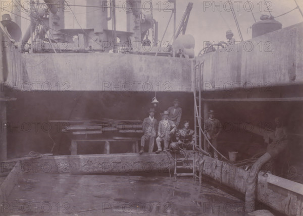 Japanese divers on hold II, photograph, A Japanese diving team poses at the edge of the hold of a dredger or suction vessel. Part of the group of individual photographs regarding the construction of the port of Belawan on the coast off Medan, Sumatra 1921-1922., inscription: 'Japanese divers on hold II', Belawan, 1921 - 1922, photographer, Sumatra, paper, daylight gelatin silver print, height 144 mm x width 198 mm, dredge, dredging machine, Japanese, deep-sea diving, economic history, overseas history, traffic and transport, infrastructure, harbor, dredging, Period, A dimly lit ship interior with a large open hold and concrete-like walls, showing a group of workers standing and sitting on wooden planks and ladders; heavy machinery, winches, ropes, pipes, and a flexible hose run along the edges, and a diving helmet and associated gear rest near the ladder; a hanging lamp is suspended above the group, and additional crew members are visible on the upper deck beside large cylindrical equi...