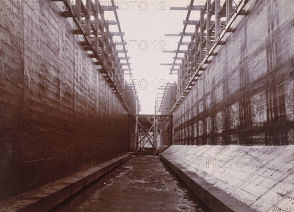 In the construction pit: ready to float, Belawan Ocean Port, photograph, Construction pit with caissons. Numbered: 65. Part of the group of loose photographs regarding the construction of the port of Belawan on the coast off Medan, Sumatra 1921-1922., date: '3, 10-21', inscription: 'No. 65, In the construction pit: ready to float', Belawan, 1921-10-03, photographer, Sumatra, paper, daylight gelatin silver print, height 160 mm x width 220 mm, harbour, building-site, economic history, overseas history, traffic and transport, infrastructure, harbor, Period, 1921 - 1922, A narrow, long concrete channel flanked by tall, textured walls with visible seams and streaks, wooden scaffolding and horizontal beams attached along the top edges on both sides, a muddy floor with shallow water and debris, stepped ledges running along the base of the walls, a framed wooden support structure spanning the channel in the distance, and an overcast sky visible above the open top.