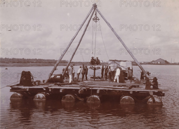 Boiling rig in the channel, Belawan Ocean Harbor, photograph, Floating raft with a drilling rig, at work in the channel of the new harbor, October 1921. Numbered: 67. Part of the group of loose photographs regarding the construction of the port of Belawan on the coast off Medan, Sumatra 1921-1922., inscription: 'No 67 Belawan Ocean Harbor, Boiling rig in the channel', date: '5, 10-21', Belawan, 1921-10-05, photographer, Sumatra, paper, daylight gelatin silver print, height 160 mm x width 220 mm, harbor, building-site, economic history, overseas history, traffic and transport, infrastructure, Period, 1921 - 1922, A large wooden raft floats on calm water with cylindrical flotation drums visible along its underside and simple mechanical winches mounted near its corners, a tall tripod of rough wooden poles rises from the raft center supporting a pulley and hanging cables, and several men in varied clothing stand and sit on the raft around a suspended platform while one man in light clothin...