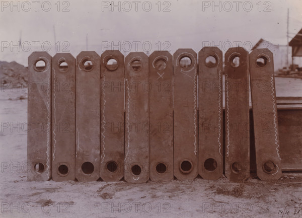 Bottle chain links, Belawan Ocean Harbor, photograph, Parts of the bucket chain of a dredging ship. Numbered: 70. Part of the group of loose photographs about the construction of the port of Belawan on the coast off Medan, Sumatra 1921-1922., inscription: 'No. 70, Bucket chain links', date: '17, 10-21', Belawan, 1921-10-17, photographer, Sumatra, paper, daylight gelatin silver print, height 160 mm x width 220 mm, harbor, building-site, economic history, overseas history, traffic and transport, infrastructure, Period, 1921 - 1922, A row of vertically oriented rectangular metal beams with octagonal tops and circular holes near the top and bottom edges, standing side by side on a sandy surface; the beams show worn and rusty surfaces with chalk-like wavy lines and circular marks on some faces; faint background elements include a distant low horizon, indistinct structures, and utility poles under a pale sky.