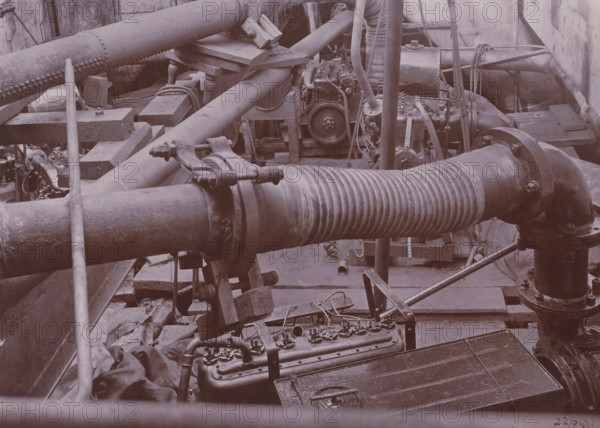Image of SS Altenburg at Singapore, Belawan Ocean Harbor, photograph, Machinery on board the SS Altenburg at Singapore. Numbered: 75. Part of the group of individual photographs regarding the construction of the port of Belawan on the coast off Medan, Sumatra 1921-1922., inscription: 'No. 75, Image of SS Altenburg at Singapore', date: '22, 9-21', Singapore, 1921-09-22, photographer, paper, daylight gelatin silver print, height 160 mm x width 220 mm, steamsailer, economic history, overseas history, traffic and transport, infrastructure, harbor, Sumatra, Belawan, Period, 1921 - 1922, A close-up view of an industrial interior filled with large metal pipes and flanged joints, a corrugated flexible pipe section running horizontally across the foreground, several heavy clamp fittings and brackets, wooden blocks and beams supporting the piping, visible bolts and nuts on flanges, coiled ropes and wire cables draped over structures, a mechanical assembly with a wheel and connected hoses and wir...
