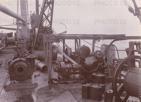 Image of SS Altenburg at Singapore, Belawan Ocean Harbor, photograph, Machinery on deck of the SS Altenburg at Singapore. Numbered: 76. Part of the group of individual photographs regarding the construction of the port of Belawan on the coast off Medan, Sumatra 1921-1922., date: '23, 9-21', inscription: 'No. 76, Image of SS Altenburg at Singapore', Singapore, 1921-09-23, photographer, paper, daylight gelatin silver print, height 160 mm x width 220 mm, steamsailer, economic history, overseas history, traffic and transport, infrastructure, harbor, Sumatra, Belawan, Period, 1921 - 1922, Machinery and heavy metal components fill the deck, including large cylindrical and drum-shaped pieces, pulleys, belts, and pipes arranged among wooden beams and metal supports, with ropes and chains draped across the structure; a lattice metal crane or derrick rises near the center, flanked by vertical columns and mounted equipment, while various fittings, flanges, and housings rest on the deck surface th...
