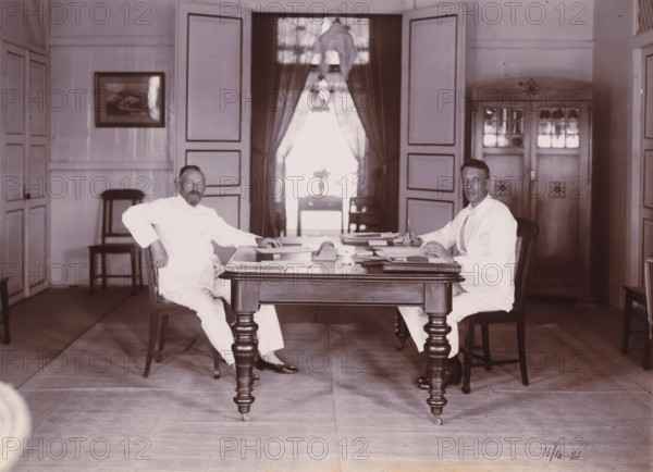 Directors of Grotius Company, Belawan Ocean Port, photograph, Interior with the two directors of the Grotius Contracting Company, sitting behind a work table. Left Laurens de Groot, right probably PJF van Buuren, October 1921. Numbered: 82. Part of the group of loose photographs concerning the construction of the port of Belawan on the coast off Medan, Sumatra 1921-1922., date: '16, 10-21', inscription: 'No 82, Directeuren Mij Grotius', Belawan, 1921-10-16, photographer, Sumatra, paper, daylight gelatin silver print, height 160 mm x width 220 mm, Person, Laurens de Groot, historical persons, office (, administration), director, boss (in modern industry), economic history, overseas history, traffic and transport, infrastructure, harbor, Period, 1921 - 1922, Two men in light-colored suits sit opposite each other at a wooden table with turned legs, papers and writing materials spread across its surface. The man on the left leans back slightly with one arm resting on the chair back and the...