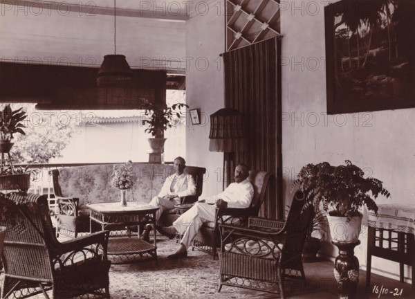 Directors of the Grotius Contracting Company, Oceaanhaven Belawan, photograph, Interior of a house with the two directors of the Grotius Contracting Company, Laurens de Groot on the right, probably PJF van Buuren on the left, October 1921. Untitled and unnumbered. Part of the group of individual photographs concerning the construction of the port of Belawan on the coast off Medan, Sumatra 1921-1922., date: '16, 10-21', Belawan, 1921-10-16, photographer, Sumatra, paper, daylight gelatin silver print, height 160 mm x width 220 mm, Person, Laurens de Groot, office (, administration), director, boss (in modern industry), economic history, overseas history, traffic and transport, infrastructure, harbor, Period, 1921 - 1922, A covered porch or veranda with wicker furniture including chairs, a round table, and a low ottoman, patterned upholstery on a settee, potted plants on stands and on the floor, two men seated in armchairs wearing light-colored clothing, a hanging fringed lampshade and a ...