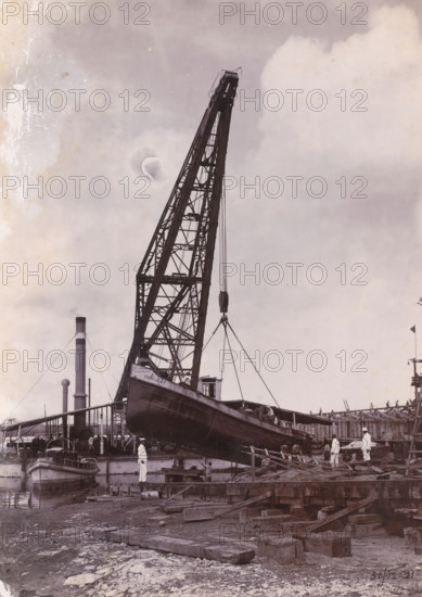 Laying out the tugboat 'Cornelis' to be repaired, Oceaanhaven Belawan, photograph, A tugboat is being lifted out of the water for repair with a crane. Numbered: 105. Part of the group of loose photographs concerning the construction of the port of Belawan on the coast off Medan, Sumatra 1921-1922., inscription: 'No 105 Ocean Port Belawan, Laying down of the tugboat 'Cornelis' to be repaired', date: '31, 12-21', Belawan, 1921-12-31, photographer, Sumatra, paper, daylight gelatin silver print, height 220 mm x width 160 mm, harbour, building-site, dock crane, repairing, caulking, careening (, ship), tug, pusher tug, economic history, overseas history, traffic and transport, infrastructure, harbor, dredging, Period, 1921 - 1922, A large industrial crane with a lattice boom towers upward, its cables suspending a wooden vessel that is angled over a dock; several men in light-colored work clothes stand on the dock and near the hull, and a small boat is moored alongside the platform; in the ba...