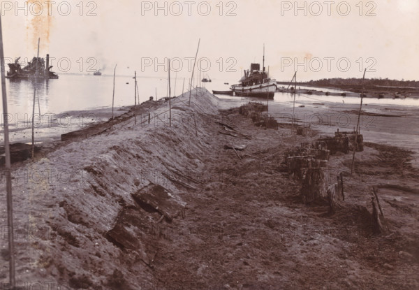 Dumping SS Madjoe (sand loading) 1st work, Belawan Ocean Port, photograph, Dredging and pumping work by the SS Madjoe at the construction pit. Numbered: 133. Part of the group of loose photographs regarding the construction of the port of Belawan on the coast off Medan, Sumatra 1921-1922., inscription: 'No. 133 Ocean Port Belawan, SS Madjoe Dump (sand load) 1st work', date: '11, 3.22', Belawan, 1922-03-11, photographer, Sumatra, paper, daylight gelatin silver print, height 160 mm x width 220 mm, harbor, building-site, dredge, dredging machine, economic history, overseas history, traffic and transport, infrastructure, dredging work, Period, 1921 - 1922, A wide, low-angle shoreline scene showing a raised, narrow earthen embankment running toward the water, with thin vertical poles driven along its length and rough, muddy ground on either side scattered with broken wooden planks and stumps; a large vessel sits moored near the end of the embankment with a smaller boat alongside it, other b...