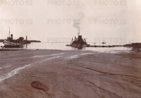 Dumping SS Madjoe (sand loading) 1st work, Belawan Ocean Port, photograph, Sand dredged by the SS Madjoe. Numbered: 134. Part of the group of loose photographs regarding the construction of the port of Belawan on the coast off Medan, Sumatra 1921-1922., inscription: 'No. 134 Ocean Port Belawan, SS Madjoe Dump (sand loading) 1st work', date: '11, 3.22', Belawan, 1922-03-11, photographer, Sumatra, paper, daylight gelatin silver print, height 160 mm x width 220 mm, harbor, building-site, dredge, dredging machine, economic history, overseas history, traffic and transport, infrastructure, dredging work, Period, 1921 - 1922, A wide view of a shoreline with wet sand in the foreground showing rippled textures and shallow water channels, a calm body of water beyond with boats and barges near the horizon, a vessel on the left with a tall mast and funnel, a group of workboats and pontoons in the center including a platform with a large wheel and a chimney emitting a plume of smoke, a small triang...