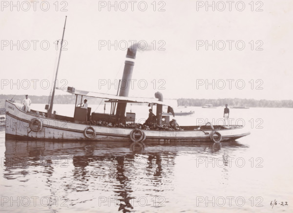 Tugboat 'Cornelis', Ocean Harbor Belawan, photograph, The tugboat 'Cornelis'. Numbered: 168. Part of the group of individual photographs regarding the construction of the harbor of Belawan on the coast off Medan, Sumatra 1921-1922., inscription: 'No 168 Tugboat 'Cornelis'', date: '6, 6-22', Belawan, 1922-06-06, photographer, Sumatra, paper, daylight gelatin silver print, height 160 mm x width 220 mm, harbor, building-site, steamship, motorship, economic history, overseas history, traffic and transport, infrastructure, dredging, Period, 1921 - 1922, A small steam-powered boat is shown floating on calm water with its reflection visible, featuring a tall smokestack emitting a plume of smoke and a corrugated canopy over the central deck area; the hull has round life rings attached along the side and a pile of coal or similar material stored under the canopy. Three men are visible on the boat, one standing near the bow wearing light-colored clothing, one seated or standing under the canopy ...