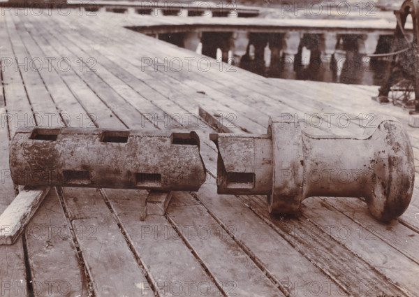 Bollard (broken), Belawan Ocean Harbor, photograph, A broken machine part (bollard). Numbered: 175b. Part of the group of loose photographs about the construction of the port of Belawan on the coast off Medan, Sumatra 1921-1922., inscription: 'No 175b Bollard (broken)', date: '30, 6-22', Belawan, 1922-06-30, photographer, Sumatra, paper, daylight gelatin silver print, height 160 mm x width 220 mm, harbor, building-site, parts of machines, economic history, overseas history, traffic and transport, infrastructure, dredging, Period, 1921 - 1922, A close view of a wooden deck with wide planks showing weathering and stains, on which two large cylindrical metal objects lie horizontally; the object on the left is a hollow, segmented cylinder with rectangular cutouts and a rough, mottled surface, while the object on the right is a heavier, flared-ended cylinder with a rounded flange and similar worn, corroded texture; wooden beams beneath the metal pieces support them, and the background shows...