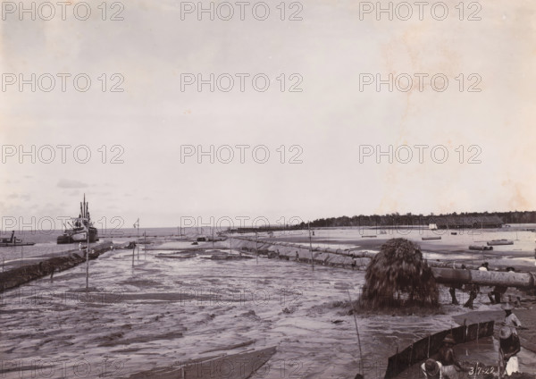 Sand loading by GG Idenburg. Height 2m + BP.), Belawan Ocean Harbour, photograph, Sand loading by the GG Idenburg. Numbered: 176. Part of the group of loose photographs regarding the construction of the port of Belawan on the coast off Medan, Sumatra 1921-1922., date: '3, 7-22', inscription: 'No 176 Sand loading by GG Idenburg. Height 2m + BP.)', Belawan, 1922-07-03, photographer, Sumatra, paper, daylight gelatin silver print, height 160 mm x width 220 mm, harbor, building-site, economic history, overseas history, traffic and transport, infrastructure, dredging, Period, 1921 - 1922, A wide shoreline scene showing wet sand and shallow water with a series of long pipes and wooden supports stretching toward the horizon, a large pile of dark material pouring from the end of a pipe into the foreground, a ship moored near a rocky jetty at the left edge, scattered small boats near the water, several small figures standing or working along the shoreline and on the jetty, low structures and a l...