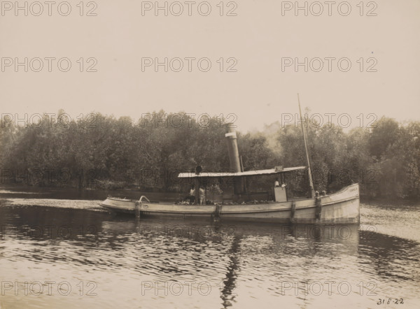 Tugboat 'Cornelis', Ocean Harbor Belawan, photograph, The tugboat 'Cornelis'. Numbered: 192. Part of the group of individual photographs regarding the construction of the harbor of Belawan on the coast off Medan, Sumatra 1921-1922., inscription: 'No. 191, Tugboat 'Cornelis'', date: '31, 8-22', Belawan, 1922-08-31, photographer, Sumatra, paper, gelatin silver print, height 160 mm x width 220 mm, harbor, building-site, tug, pusher tug, economic history, overseas history, traffic and transport, infrastructure, dredging, Period, 1921 - 1922, A weathered steam launch floats on calm water near a tree-lined riverbank, its hull showing signs of age and ropes draped over the side, a covered central cabin topped by a vertical smokestack emitting a faint plume, a small boat tied alongside, a slender mast rising toward the sky, and trees densely filling the background while ripples and reflections ripple across the water surface.