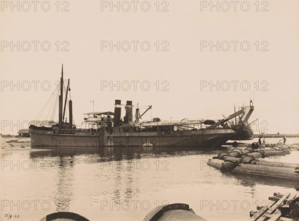 Sludge and sand dredger GG Idenburg, Ocean Harbour Belawan, photograph, The hopper dredger GG Idenburg. Numbered: 203. Part of the group of loose photographs about the construction of the harbour of Belawan on the coast off Medan, Sumatra 1921-1922., inscription: 'No. 203 Silt and sand dredger GG Idenburg', date: '15, 9-22', Belawan, 1922-09-15, photographer, Sumatra, paper, gelatin silver print, gelatin silver developing print, height 160 mm x width 220 mm, harbor, building-site, steamship, motorship, economic history, overseas history, traffic and transport, infrastructure, dredging, Period, 1921 - 1922, A large steamship sits low in calm water near a shore, its hull dark and weathered with visible seams and a ladder hanging down the side, two tall smokestacks rise amidship flanked by masts and rigging, and a deck structure with open and covered areas containing equipment and walkways. A long angled ramp or conveyor extends from the ship toward the right, meeting a series of large cy...