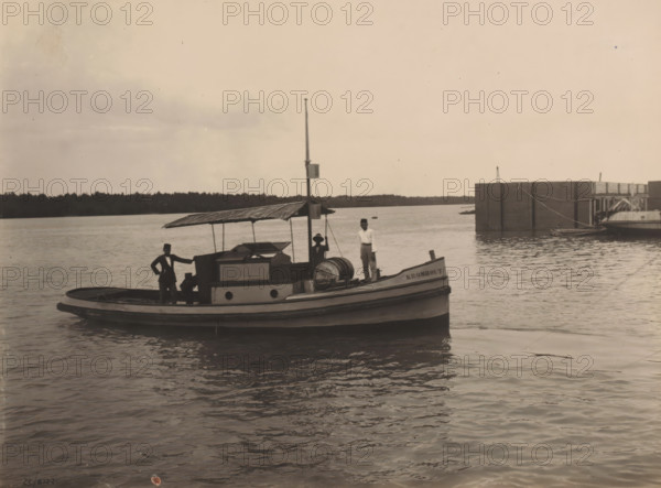 Motorboat 'Kromhout', Ocean Harbor Belawan, photograph, Motorboat 'Kromhout'. Numbered: 214. Part of the group of individual photographs regarding the construction of the harbor of Belawan on the coast off Medan, Sumatra 1921-1922., inscription: 'No. 214 Motorboat 'Kromhout'', date: '25, 9-22', Belawan, 1922-09-25, photographer, Sumatra, paper, gelatin silver print, height 160 mm x width 220 mm, harbor, building-site, steamship, motorship, economic history, overseas history, traffic and transport, infrastructure, dredging, Period, 1921 - 1922, A small wooden boat with a covered cabin sits on calm water near a larger dock structure; three men stand on the boat, two wearing hats and one holding a rope, while one man leans with a hand on the cabin roof; the boat has a rounded bow and visible deck equipment including a large barrel and a vertical mast with a small box attached; a distant treeline runs along the horizon under a mostly clear sky.