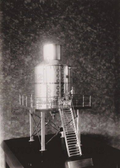 Scale model of a cooking pot, photograph, without the glass cover of the display cabinet. Part of the binder with photos of machines from NV Machinefabriek Braat in Surabaya intended for the Dutch sugar industry in Java, from the period 1917-1938., Surabaya, 1917 - 1938, photographer, Java, East Java, paper, gelatin silver print, gelatin silver print on photographic paper, height 180 mm x width 130 mm, Person, NV Machinefabriek Braat, colonial system, (non-fruit) products of plants or trees: sugarcane, machines, parts of machines, tools and appliances, economic history, overseas history, sugar industry, business history, Period, A metallic cylindrical tank with a domed top sits elevated on a square platform supported by four angled legs with cross-bracing, a narrow ladder leading up to the platform, and a railing around the platform edge; the tank surface is reflective and features a vertical row of circular openings on one side and a pair of vertical pipes and valves, with various sma...