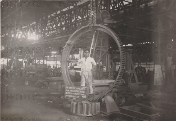 Gear ring for reed unloading device, photograph, Cast steel gear ring for a reed unloading device, manufactured in 1925 and intended for the Gempolkrep Sugar Factory. Part of the binder containing photographs of machines from NV Machinefabriek Braat in Surabaya intended for the Dutch sugar industry in Java, from the period 1917-1938., Surabaya, 1925, photographer, Java, East Java, paper, gelatin silver print, gelatin silver print on photographic paper, height 121 mm x width 173 mm, Person, NV Machinefabriek Braat, colonial system, (non-fruit) products of plants or trees: sugarcane, machines, parts of machines, tools and appliances, factory-interior, machine shop (laborers, crafts, industries, agriculture), economic history, overseas history, sugar industry, business history, Period, 1917 - 1938, A man in light-colored work clothes stands inside a large industrial workshop, his hand resting on a large circular gear that hangs from a chain; the gear has visible teeth along its outer edge...
