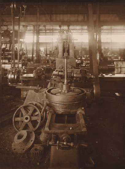 Milling machine, photograph, The first straight gear milling machine from ca. 1916. Part of the binder with photos of machines from the NV Machinefabriek Braat in Surabaya, Sukabumi, and Yogyakarta, from the period 1908-1942., Surabaya, 1919 - 1921, photographer, Java, East Java, paper, gelatin silver print, height 202 mm x width 152 mm, Person, NV Machinefabriek Braat, colonial system, factory-interior, machine shop, machines, parts of machines, tools and appliances, economic history, overseas history, business history, Period, 1908 - 1942, A sepia-toned photograph of an industrial workshop showing a large metal machine in the foreground with a vertical shaft, gears, and circular components, a set of toothed wheels and flanges resting nearby on the floor, metal rails or guides leading toward the machine, and additional blurred machines, columns, and windows in the background with light streaming through the windows.